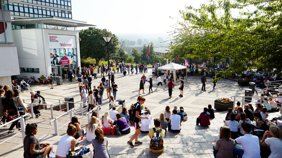 A busy outdoor plaza at a university campus with groups of people sitting on steps and walking across the square. The scene includes a white building with a large “Welcome” sign, trees providing shade, and a white canopy tent in the centre. In the background, there are more people gathered and distant hills visible under bright daylight.