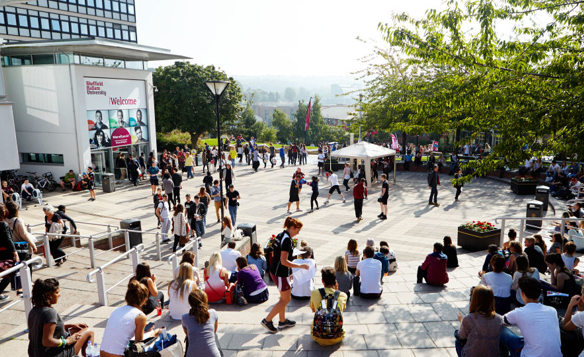 A busy outdoor plaza at a university campus with groups of people sitting on steps and walking across the square. The scene includes a white building with a large “Welcome” sign, trees providing shade, and a white canopy tent in the centre. In the background, there are more people gathered and distant hills visible under bright daylight.