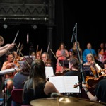 An orchestra rehearses on a theatre stage with a conductor leading from the front. String players sit with sheet music and raised bows, while a microphone stand stands at centre stage. Additional performers and musicians are visible in the background, with warm lighting focused on the ensemble and dark stage curtains behind.
