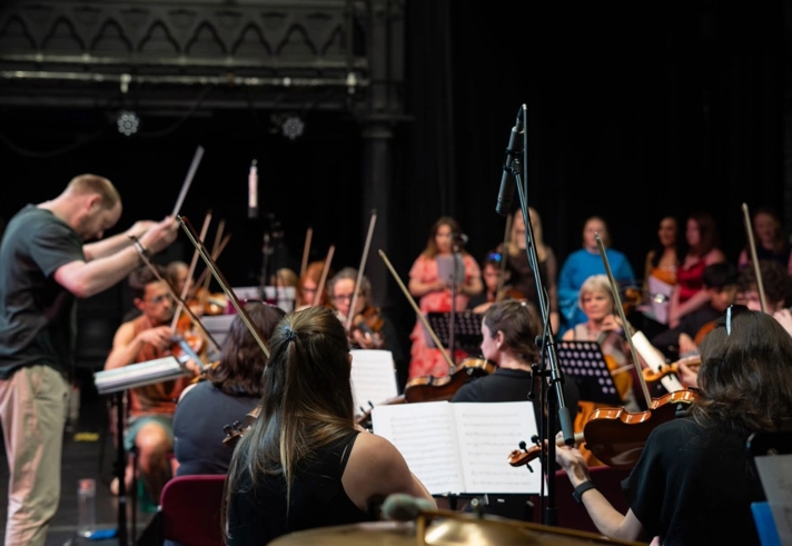 An orchestra rehearses on a theatre stage with a conductor leading from the front. String players sit with sheet music and raised bows, while a microphone stand stands at centre stage. Additional performers and musicians are visible in the background, with warm lighting focused on the ensemble and dark stage curtains behind.