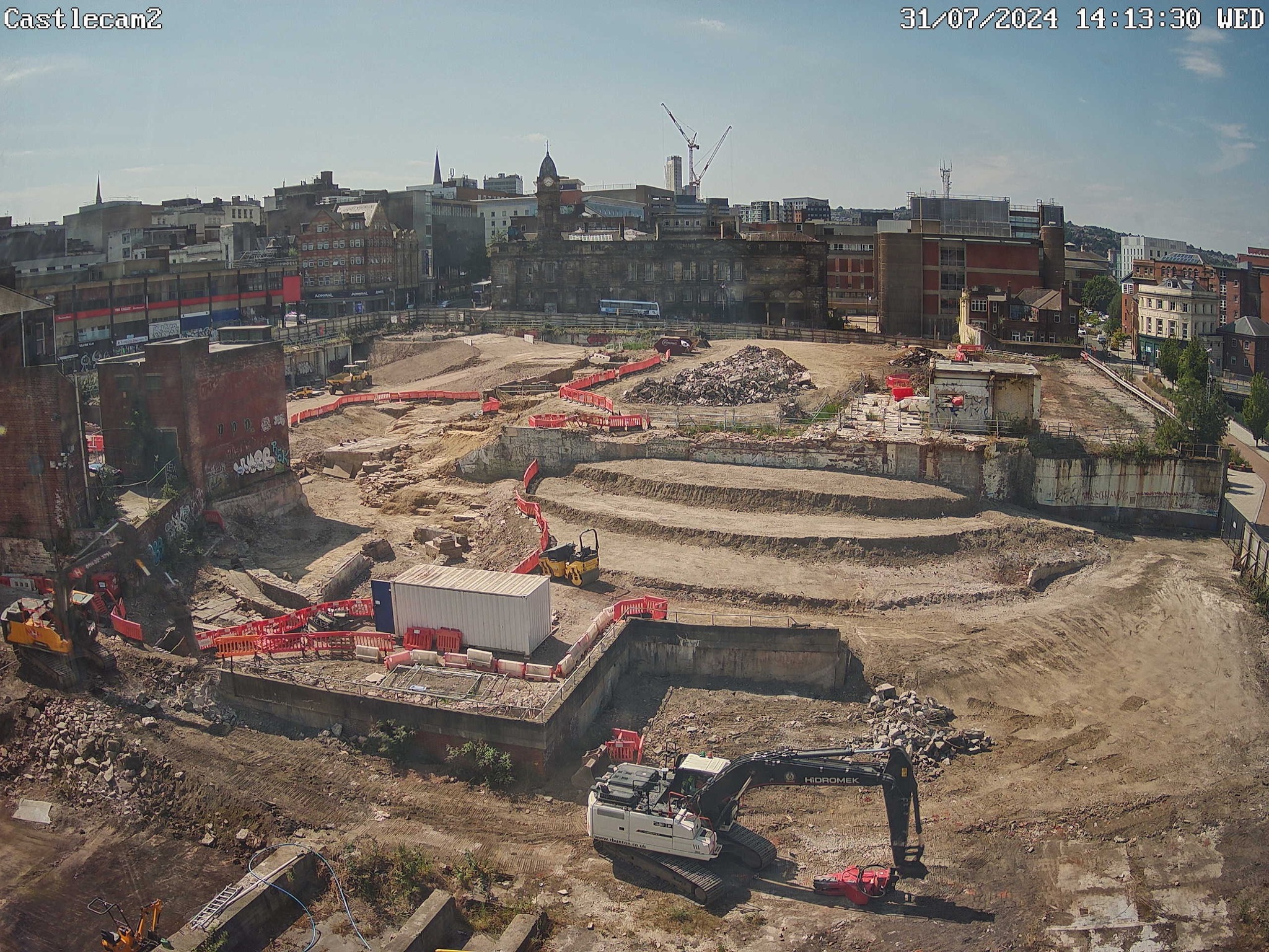 Construction site in an urban area with terraced excavation levels, machinery, and red safety barriers. Surrounding the site are older brick buildings and cranes in the background under a clear blue sky.