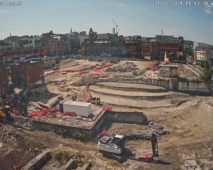 Construction site in an urban area with terraced excavation levels, machinery, and red safety barriers. Surrounding the site are older brick buildings and cranes in the background under a clear blue sky.