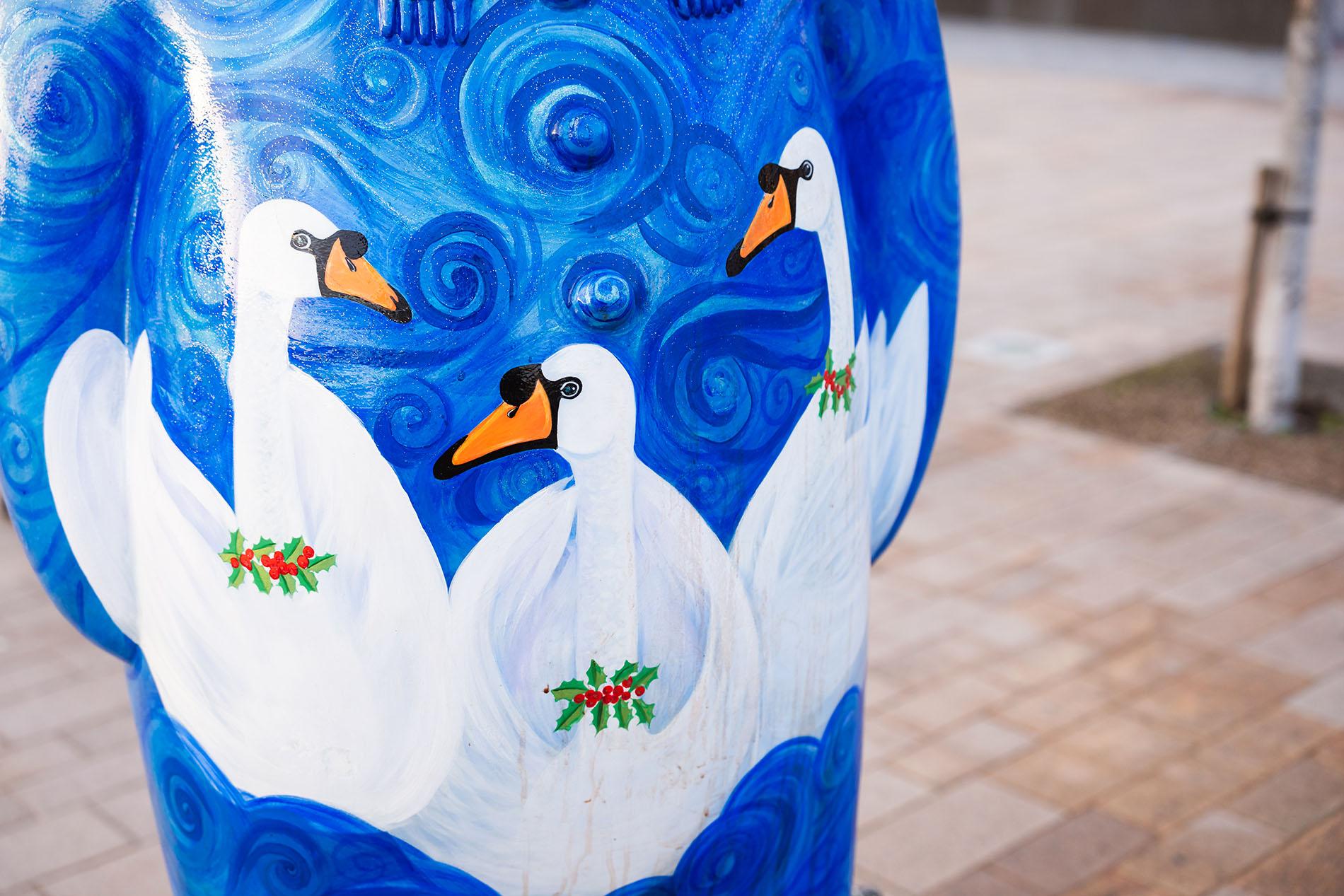 Close-up of a painted design featuring three white swans with orange beaks and holly wreaths around their necks, set against a swirling blue background. The artwork appears on a sculpted figure in an outdoor paved area.