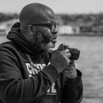 Black-and-white photograph of a man wearing glasses and a dark hoodie, seated beside a waterfront. He holds a camera in both hands and looks thoughtfully across the water. Large rocks line the shoreline in the foreground, while buildings and the distant waterfront appear softly blurred in the background, creating a calm and reflective atmosphere.