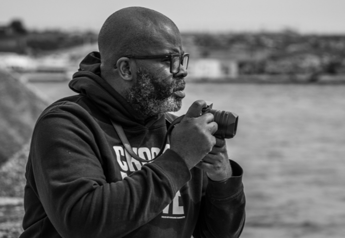 Black-and-white photograph of a man wearing glasses and a dark hoodie, seated beside a waterfront. He holds a camera in both hands and looks thoughtfully across the water. Large rocks line the shoreline in the foreground, while buildings and the distant waterfront appear softly blurred in the background, creating a calm and reflective atmosphere.