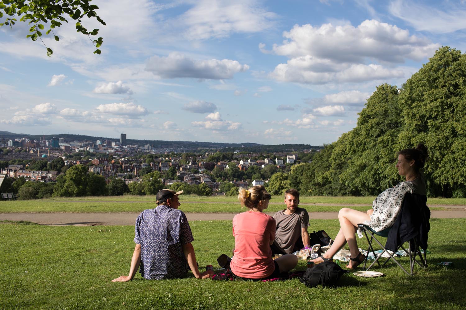 A group of four people, sat in Meersbrook Park, enjoying a sunny day. One is sat in a folding chair and the other three are sat on the grass.