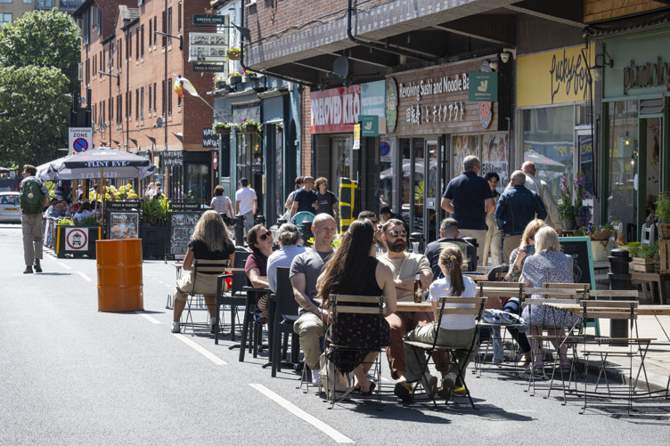 People are seated at outdoor tables on a closed-off city street, enjoying food and drinks in sunny weather. The street is lined with shops and restaurants, including signs for a noodle bar and a café. A bright orange barrel and potted plants decorate the area, while pedestrians walk along the street in the background. The scene conveys a lively, social atmosphere in an urban setting.