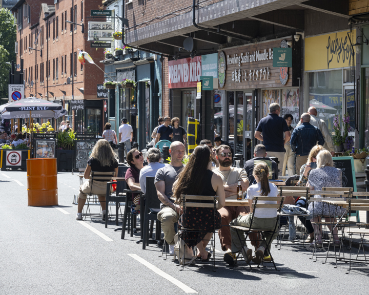 People are seated at outdoor tables on a closed-off city street, enjoying food and drinks in sunny weather. The street is lined with shops and restaurants, including signs for a noodle bar and a café. A bright orange barrel and potted plants decorate the area, while pedestrians walk along the street in the background. The scene conveys a lively, social atmosphere in an urban setting.