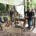 People working at wooden benches in an outdoor woodland workshop, using hand tools under a simple canopy surrounded by trees.