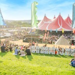 Large outdoor music festival with crowds gathered on a grassy hill and around a big red-and-white striped tent. Colorful vertical flags line the area, and smaller tents and stalls are visible in the background under a bright blue sky.
