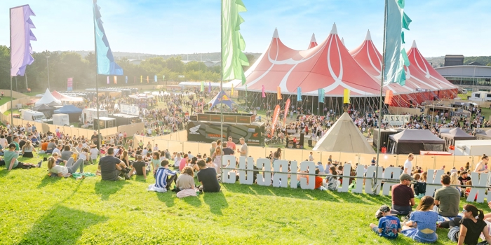 Large outdoor music festival with crowds gathered on a grassy hill and around a big red-and-white striped tent. Colorful vertical flags line the area, and smaller tents and stalls are visible in the background under a bright blue sky.