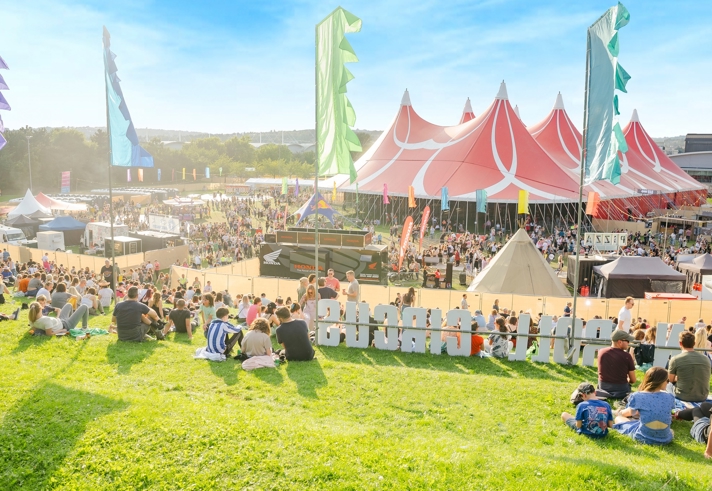 Large outdoor music festival with crowds gathered on a grassy hill and around a big red-and-white striped tent. Colorful vertical flags line the area, and smaller tents and stalls are visible in the background under a bright blue sky.