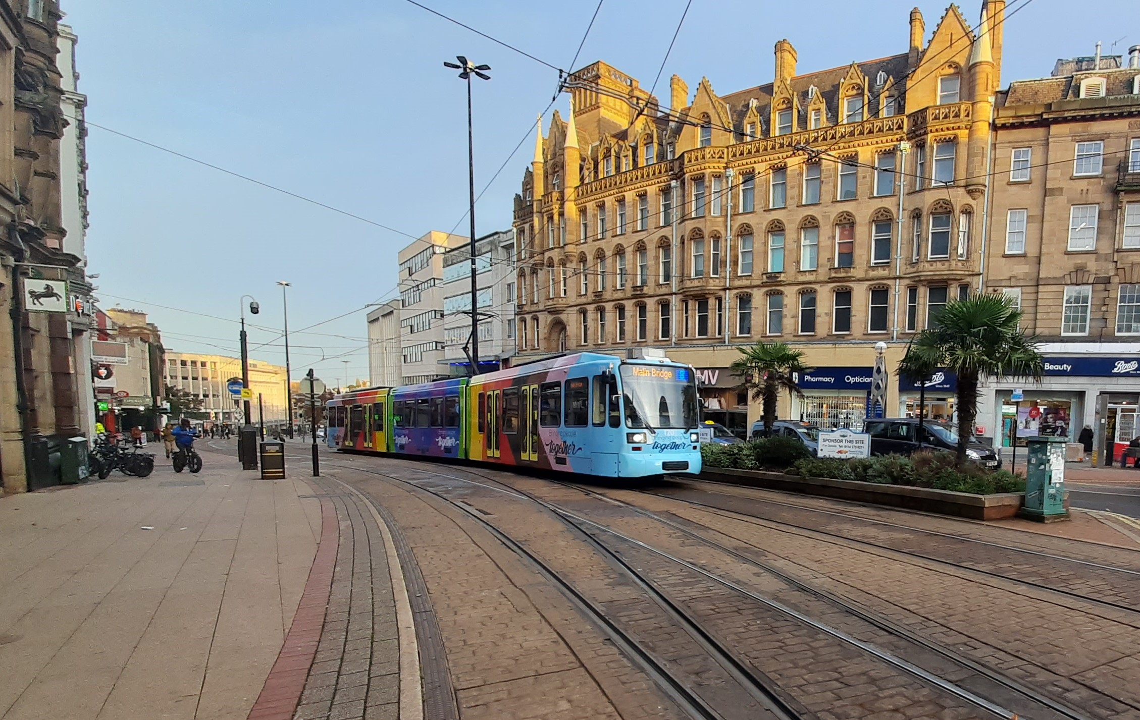 Street scene featuring a colorful tram traveling along tracks in a city center. The tram is painted in bright blue and multicolored designs. Surrounding architecture includes ornate historic buildings with large windows and decorative stonework. Palm trees and planters line the street, and pedestrians are visible in the background under a clear blue sky.