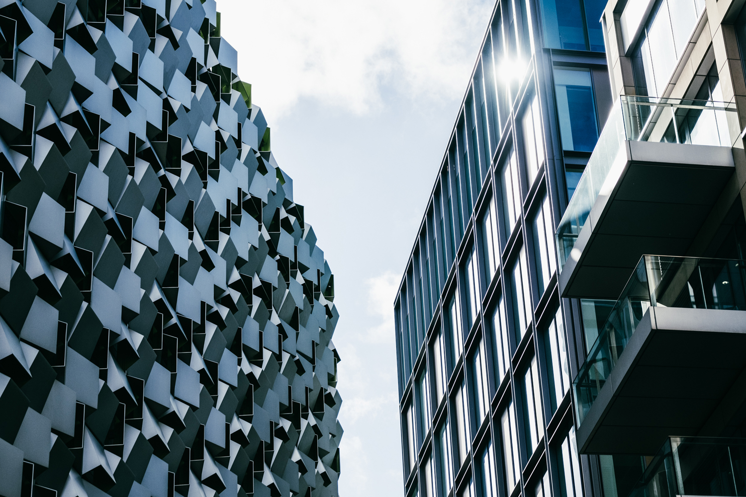 Two modern buildings with contrasting architectural styles viewed from street level against a partly cloudy sky. The building on the left features an irregular, geometric façade with protruding panels creating a textured pattern, while the building on the right has a sleek glass exterior with reflective windows and balconies. Sunlight reflects off the glass panels, adding brightness to the scene.