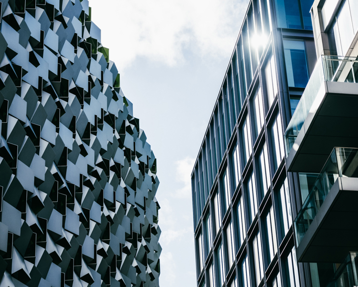 Two modern buildings with contrasting architectural styles viewed from street level against a partly cloudy sky. The building on the left features an irregular, geometric façade with protruding panels creating a textured pattern, while the building on the right has a sleek glass exterior with reflective windows and balconies. Sunlight reflects off the glass panels, adding brightness to the scene.