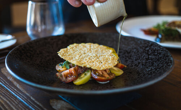 A close-up of a dark textured plate containing a colorful dish of sliced heirloom tomatoes topped with a large, crisp parmesan tuile. A hand is pouring a light green sauce or dressing from a small white jug onto the dish. In the background, there is a glass of water and another plate with food, slightly out of focus, on a wooden table.