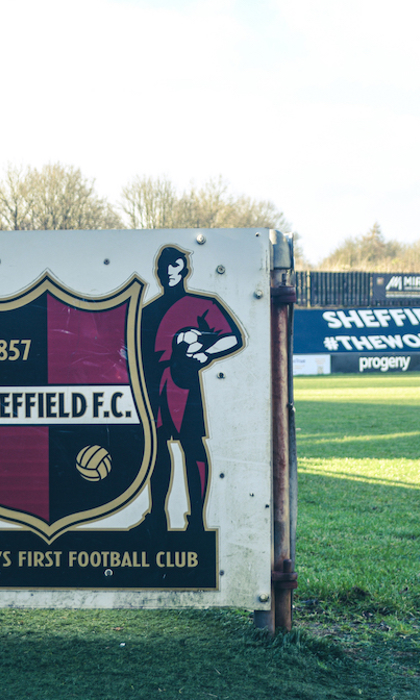 Signboard featuring the Sheffield F.C. crest with text ‘1857 Sheffield F.C.’ and ‘The World’s First Football Club,’ flanked by illustrations of two footballers. In the background, a football pitch and advertising boards display ‘Sheffield FC #TheWorldsFirst.’ Trees and sky are visible beyond the stands.