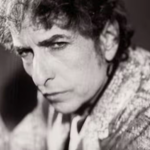 Black-and-white close-up portrait of a person with curly hair, shown from the shoulders up and leaning slightly forward. The face is turned towards the camera with a direct gaze. The person wears a patterned jacket or coat. The background is blurred and dark, drawing attention to the facial features and textured clothing.