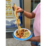 A person wearing a pink polo shirt holds a bowl of Chinese hand-pulled noodles topped with beef, bean sprouts and chilli flakes outside a restaurant. With chopsticks, they lift a long, wide ribbon of noodle stretching high above the bowl, coated in chilli oil and seasoning. The restaurant window and signage are visible in the background.