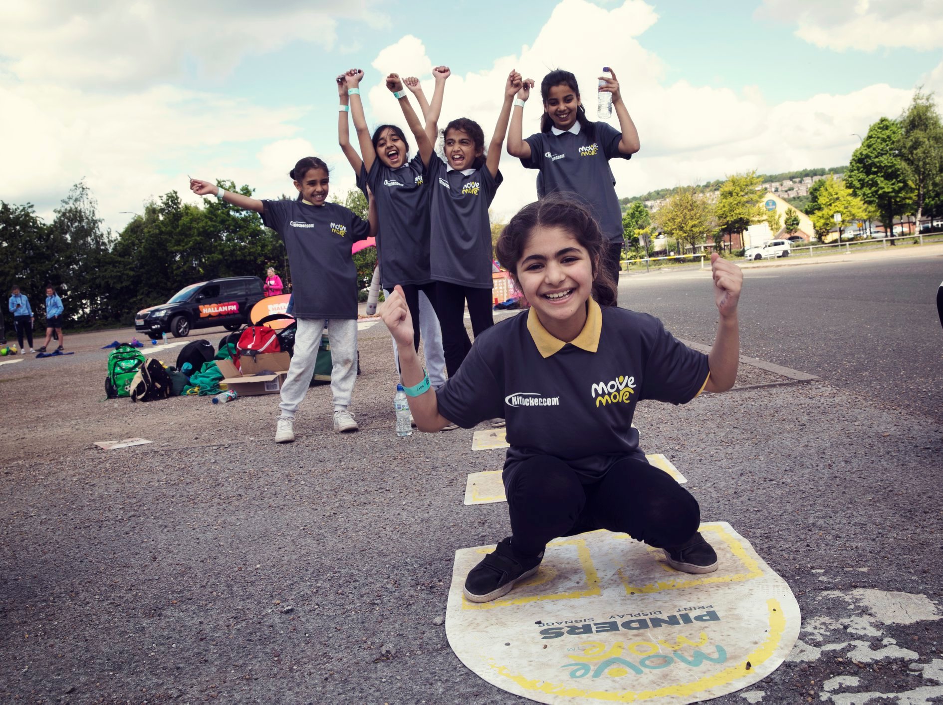 A group of children wearing matching dark gray “Move More” t-shirts participate in an outdoor activity on a paved area. One child is crouching in the foreground on a circular marker with text, while others stand behind with arms raised in celebratory poses. Sports bags and equipment are visible on the ground, and trees and parked cars can be seen in the background under a partly cloudy sky.