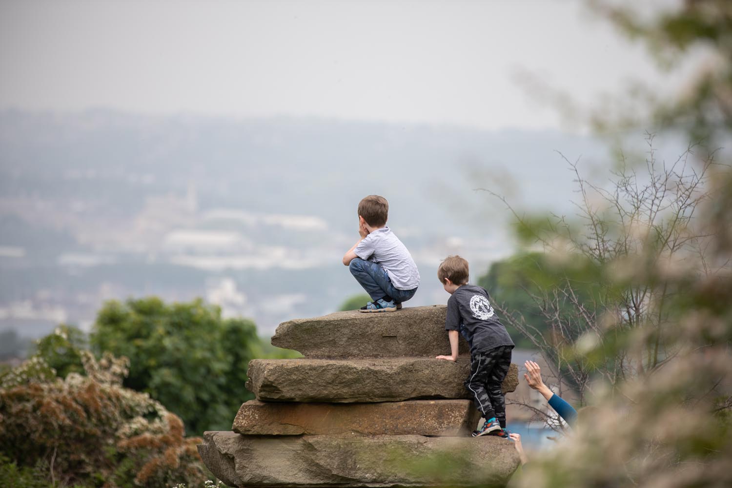 Two children are climbing and sitting on large stacked stone slabs in a natural outdoor setting. The background shows a hazy view of a distant town or city with trees and greenery in the foreground.