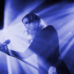 Black-and-white concert photograph of a performer standing at a microphone on stage. Bright beams of light radiate from behind, creating a dramatic silhouette effect and illuminating long hair and the microphone stand. The image has strong contrast, with stage lighting filling the background and the performer positioned in the centre foreground.