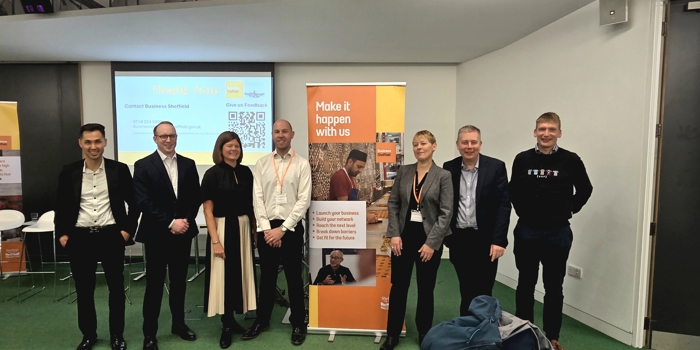 Group of seven people standing indoors in front of a presentation screen and a tall orange banner. The banner reads ‘Make it happen with us’ and includes text about business support along with an image of a person working on a laptop. The presentation screen behind them displays a thank-you message with contact details and a QR code. The setting appears to be a professional event or networking session with green carpet flooring and white walls.