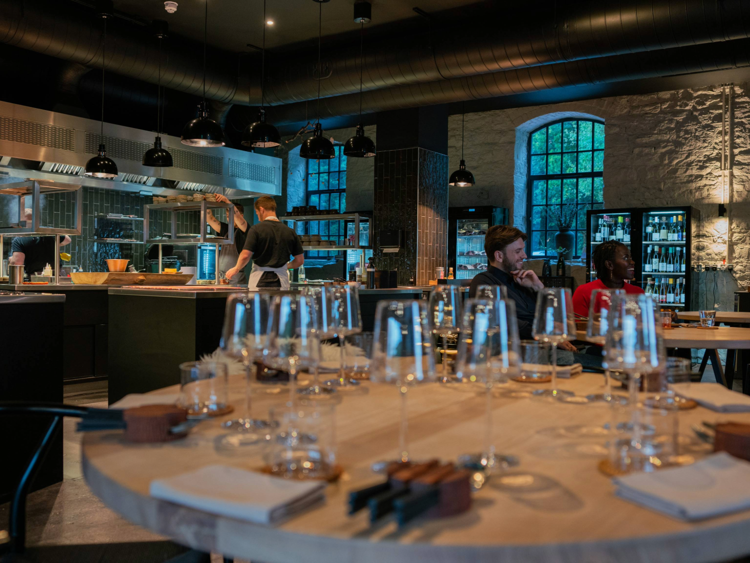A modern, stylish restaurant interior featuring an open kitchen with a chef at work. In the foreground, a round wooden table is set with multiple empty wine glasses, folded napkins, and cutlery. The space has exposed stone walls, large arched windows, and shelves stocked with bottles. Black pendant lights hang from the ceiling, and the atmosphere is warm and contemporary.
