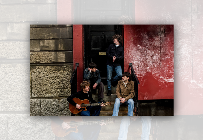 Four musicians gather on the steps outside a building with a black doorway and red-painted wall. One person sits holding an acoustic guitar while three others sit or stand on the steps nearby. The scene is framed by textured stonework on one side and weathered red paint on the other, creating an informal band portrait in an urban setting.