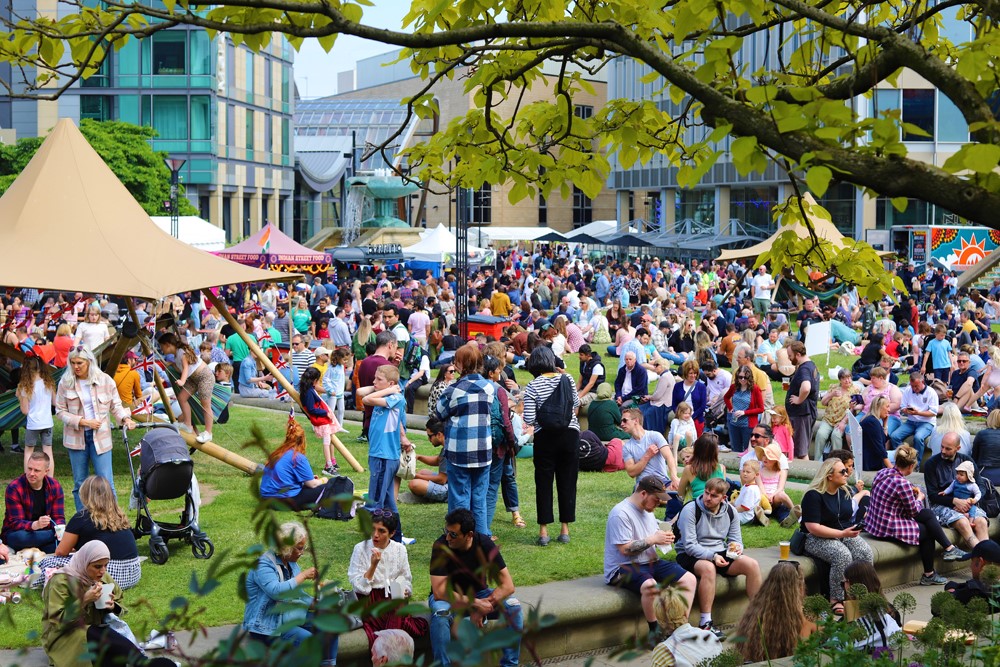 A leaf framed view as a huge crowd of people sit and stand around in a public square surrounded by street food stands on a sunny day 