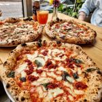Three Neapolitan-style pizzas on a wooden table near a window, each topped with different ingredients, with a small vase of flowers and a drink beside them.