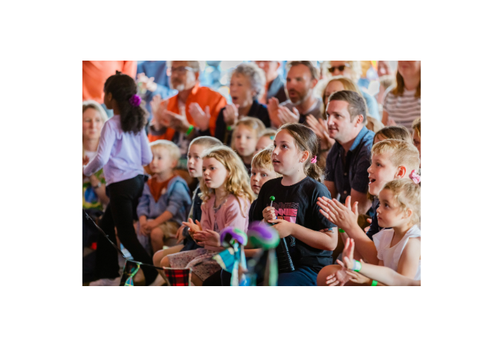 Children and adults sit closely together in an audience at a family-friendly indoor event. Several children in the foreground watch a performance, some clapping, while one child holds a green lollipop. A person stands at the left edge facing away from the camera. The background shows a packed crowd, many applauding. Bright, warm lighting and shallow focus emphasise the children, with the audience softly blurred behind them.