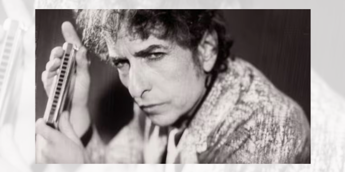 Black-and-white close-up portrait of a person with curly hair, shown from the shoulders up and leaning slightly forward. The face is turned towards the camera with a direct gaze. The person wears a patterned jacket or coat. The background is blurred and dark, drawing attention to the facial features and textured clothing.
