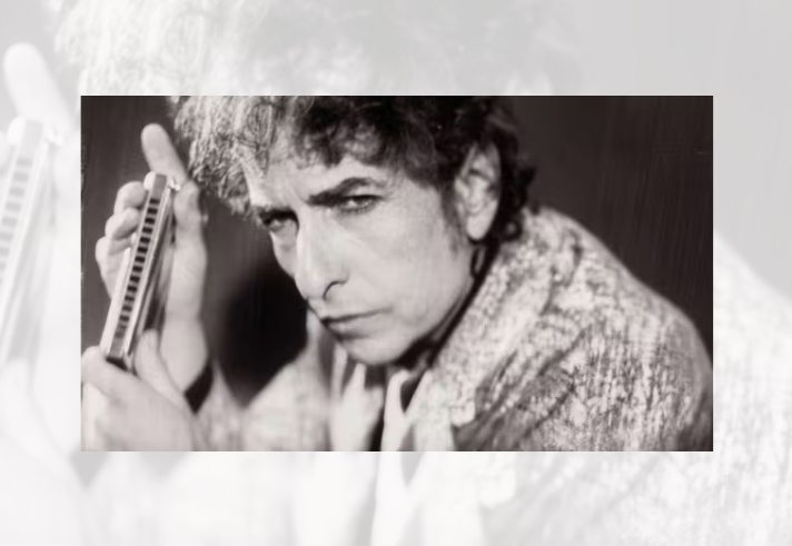 Black-and-white close-up portrait of a person with curly hair, shown from the shoulders up and leaning slightly forward. The face is turned towards the camera with a direct gaze. The person wears a patterned jacket or coat. The background is blurred and dark, drawing attention to the facial features and textured clothing.