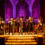 A five piece band perform on a stage, surrounded by candles. 