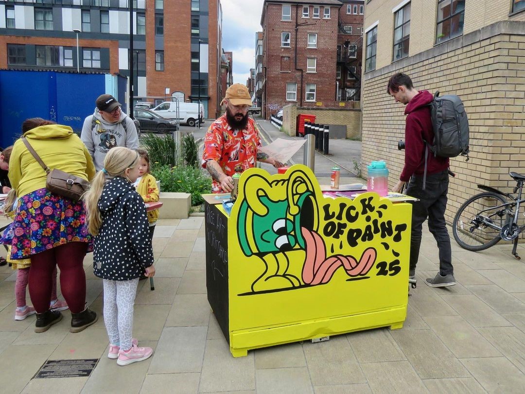 Outdoor street scene with a small art stall featuring a bright yellow counter painted with a cartoon character and the words ‘LICK OF PAINT 25.’ Several people, including children, are gathered around the stall. The background shows modern brick buildings, a bicycle leaning against a wall, and a paved walkway.