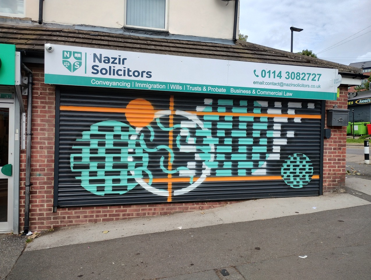 A solicitor’s office with a closed shutter painted in a modern geometric mural. The design features teal and white checkerboard patterns, two circular shapes, and an orange vertical and horizontal line intersecting at the center. Above the shutter is a sign reading “Nazir Solicitors” with contact details and services listed, including conveyancing, immigration, wills, trusts, probate, and business law.