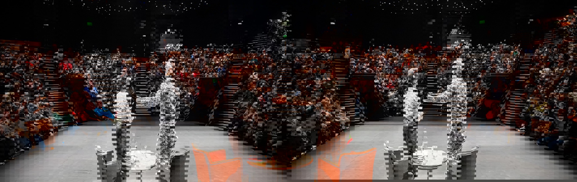 Two speakers standing on a stage in front of a full audience inside a theatre with star‑like ceiling lights.