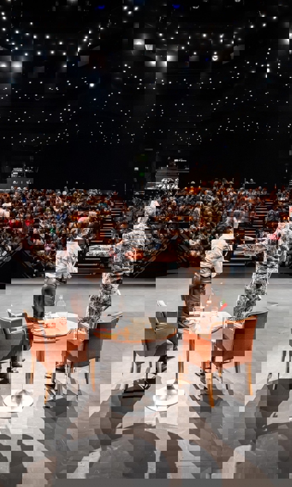 Two speakers standing on a stage in front of a full audience inside a theatre with star‑like ceiling lights.