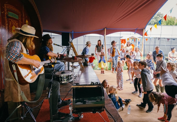 A lively scene under a large tent where two musicians are performing on a small stage. One musician is playing an acoustic guitar while the other is seated at a drum set. In front of the stage, a group of children and adults are gathered, some dancing and others watching the performance. The area is decorated with colourful bunting, and the background shows outdoor structures and greenery, suggesting a festival or community event.