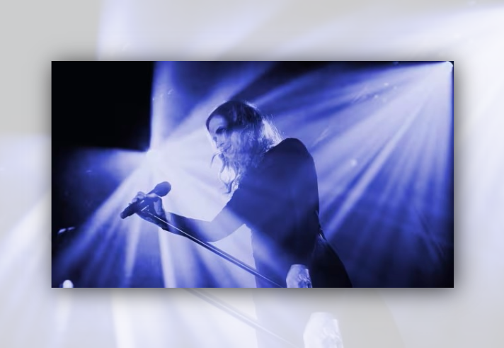 Black-and-white concert photograph of a performer standing at a microphone on stage. Bright beams of light radiate from behind, creating a dramatic silhouette effect and illuminating long hair and the microphone stand. The image has strong contrast, with stage lighting filling the background and the performer positioned in the centre foreground.