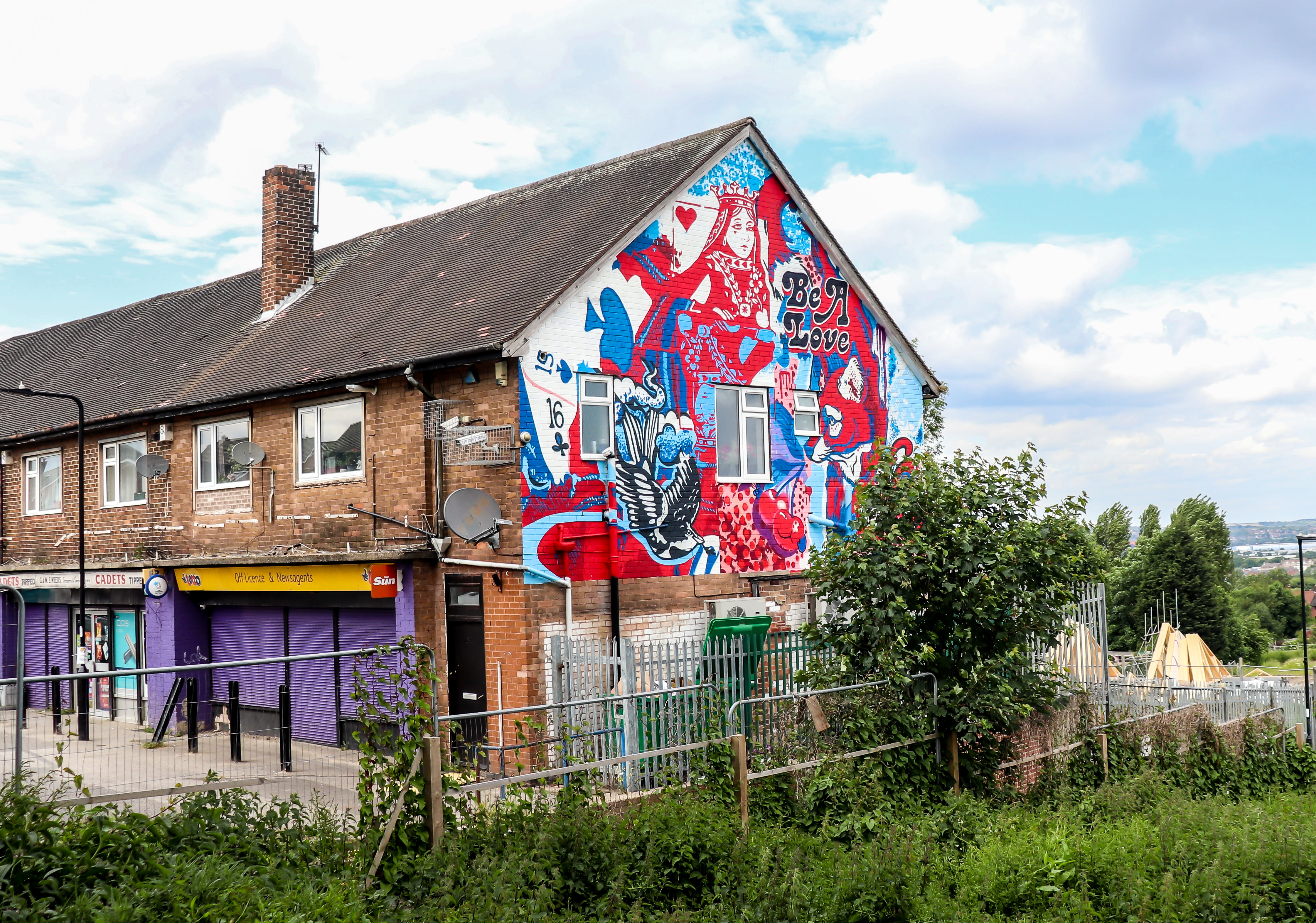 Street mural on the side of a brick building featuring bold red, blue, and white artwork with playing card motifs, a large Queen of Hearts figure, and the words ‘Be A Love.’ The mural includes hearts, flowers, and a bird design. Below the mural are shopfronts with purple shutters and signage, and greenery in the foreground.