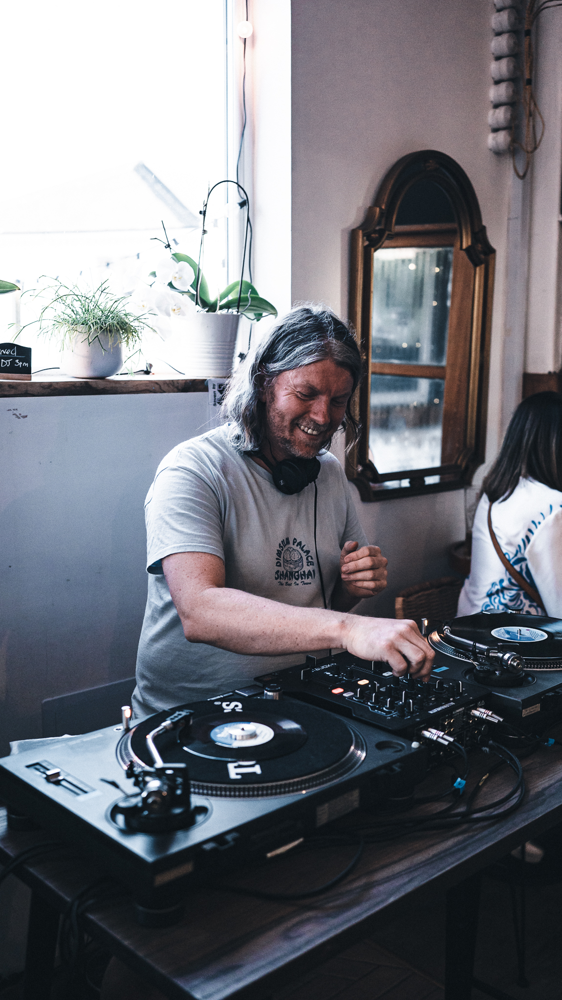 A DJ operating turntables and a mixer inside a bright room, with plants and a decorative mirror in the background.