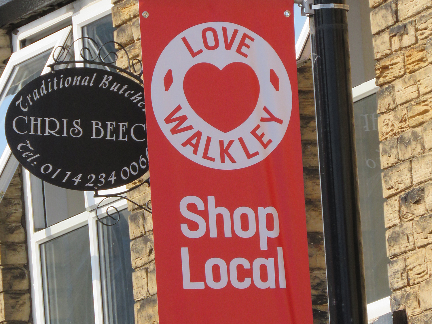 Close-up of a red banner attached to a lamppost with the words “LOVE WALKLEY” and “Shop Local” in white text, featuring a heart symbol. Next to it is a black oval sign for a traditional butcher shop reading “Chris Beec…” with a phone number partially visible. The background shows a stone building with a window.