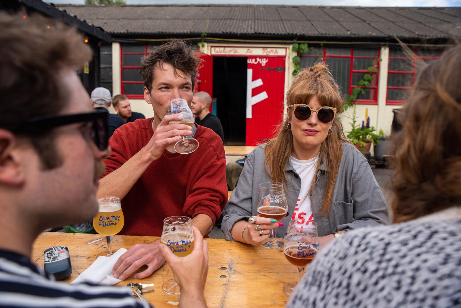 People sitting around a wooden outdoor table at a brewery, holding glasses of beer. The background shows a rustic building with red-framed windows and a bright red door featuring a white design. Plants and string lights add to the casual, social atmosphere.