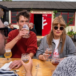 People sitting around a wooden outdoor table at a brewery, holding glasses of beer. The background shows a rustic building with red-framed windows and a bright red door featuring a white design. Plants and string lights add to the casual, social atmosphere.