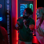 Two people standing near a neon-lit arcade game with glowing buttons and a bright display. Both are holding drinks in cocktail glasses, one orange and one clear, while watching the game. The machine features colourful lights and text reading “Green buzzers are great.” The setting is vibrant and modern, with a lively atmosphere and illuminated signage in the background.