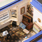 Overhead view of a detailed miniature living room styled like the 1980s or early 1990s. The tiny room features a television displaying a retro video game, shelves filled with cassette tapes, a radio, and vintage home computer equipment including a Commodore 64 box and accessories scattered on the carpet. Open bay windows, patterned flooring and carefully crafted furnishings create a nostalgic scene celebrating early home gaming culture.
