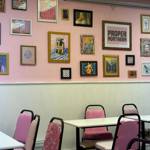 A pastel pink café-style room with white tables and pink chairs, featuring a gallery wall of eclectic framed artwork and a large “Proper Northern” print.