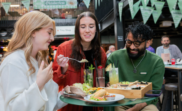 Three people seated at a round green table in an indoor food hall, sharing a meal. The table holds plates of tacos, salad, bread, and a wooden tray with assorted dishes. There are also tall glasses of colourful drinks garnished with fruit and herbs. In the background, green triangular banners hang overhead, and signs for “Oktoberfest” and “Baity” are visible, creating a lively, casual dining atmosphere.